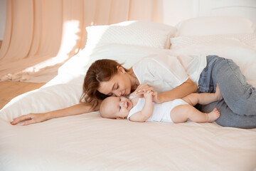 Obraz na płótnie Canvas Happy young mother with baby at home in white room, mom and baby are lying, kissing the baby at home on the bed in the bedroom