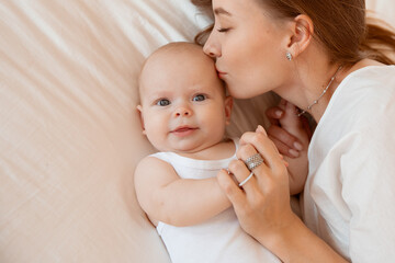 Happy young mother with baby at home in white room, mom and baby are lying, kissing the baby at...