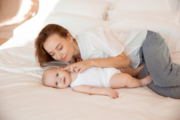 Happy young mother with baby at home in white room, mom and baby are lying, kissing the baby at home on the bed in the bedroom