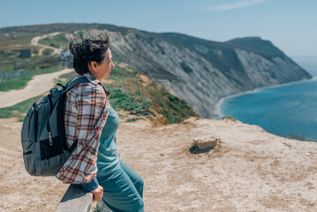 an adult woman with a backpack went hiking in the mountains near the sea in summer, a spring sunny day for health. pensioner, sat down to rest on a bench on top of a mountain, admires the sea, ocean