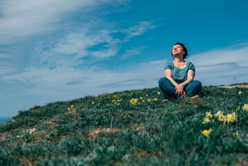 Naklejka na ściany i meble an adult woman, pensioner sits on the grass, legs crossed in a lotus position barefoot with a smile on her face on a sunny day on a mountain against the backdrop of the sea in summer, spring