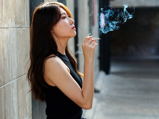 Portrait of beautiful Chinese girl with black long hair in black sexy sundress leaning against wall and looking away with smoking cigarette in hand, smoke coming out from mouth.