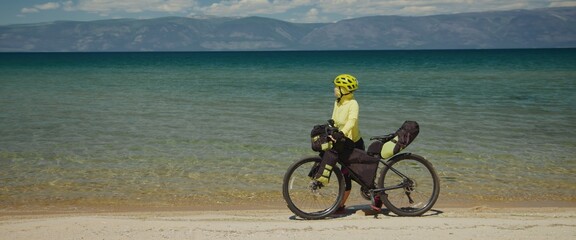 Fototapeta premium The girl towing gravel bike on the beach against the sea background. The woman travels on a bicycle. The cycle in modern style sportswear, helmet and sport glasses in green black colors.