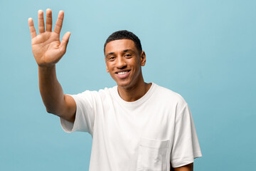 Friendly young african-american guy wearing white casual t-shirt waving hand hi isolated on blue background, happy millennial man looking at camera welcoming you, greeting gesture