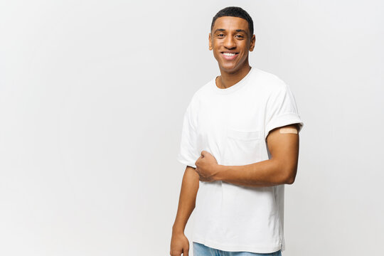 Portrait of happy young African American guy showing arm with patch-aid after coronavirus vaccine injection isolated on blue background. Man protect himself against viral diseases