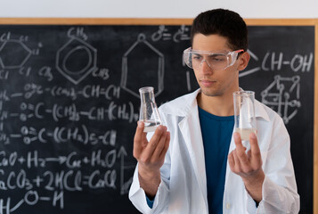 A young Arab male chemistry teacher stands against the background of a chalk board with different formulas, dressed in a white coat and goggles, holding two different test tubes with reagents.