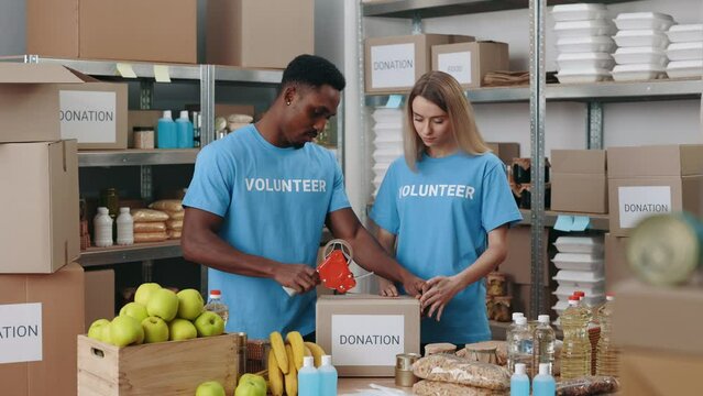 Two diverse volunteers packing and sticking cardboard boxes at food bank warehouse. Young african american man and caucasian woman helping people in need.