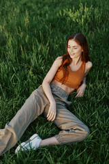 Naklejka na ściany i meble A young, happy woman lies on the green grass in the park and relaxes in the sunset summer light. The concept of health and harmony with nature