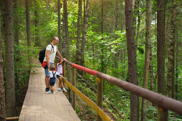 Obraz na płótnie Canvas Dad and daughters are standing on old wooden footbridge in woods among trees. Hiking with whole family with small kid. Traveling through ecological trail. Man with hiking backpack holds girls by hands