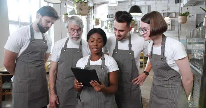 African American ana Caucasian team of cooks standing in restaurant, watching something on tablet device and talking. Planning menu. Multi-ethnic waiters using computer in cafe. Colleagues in bar.