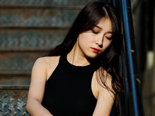 Graceful young woman with long black hair sitting on ground and leaning on the wall with eyes closed in sunny day. Outdoor portrait of cool Chinese girl in sexy black sundress, summer concept.