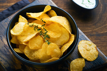 Potato chips. Fast food. Crispy potato chips ceramic black bowl with sour cream sauce and onions in wooden stand on old kitchen table wooden background. American tradition. Hot BBQ. Top view.
