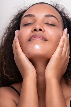 Young Hispanic woman applying cream of face with closed eyes