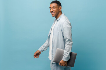 Friendly young african-american male student in casual shirt carrying laptop pc isolated on blue background, smiling multiracial freelancer man looking at the camera