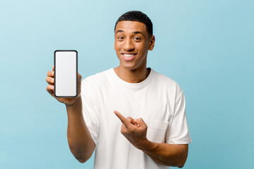 Smiling African-American young guy points index finger at empty smartphone screen isolated on blue...
