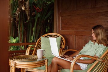 Young beautiful woman working with a laptop and a cup of coffee on the porch or veranda of a wooden summer house or bungalow