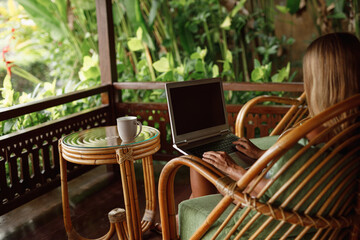 Young beautiful woman working with a laptop and a cup of coffee on the porch or veranda of a wooden summer house or bungalow