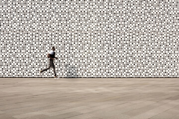 Unrecognizable male athlete running along sidewalk on background of modern art center with white perforated facade, blurred motion