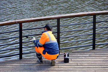 Worker in uniform paints the metal railing on a lake or river embankment. Repair works in spring...