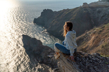 Woman tourist enjoying the sunset over the sea mountain landscape. Sits outdoors on a rock above the sea. She is wearing jeans and a blue hoodie. Healthy lifestyle, harmony and meditation