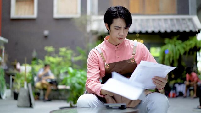 Bored anxiety Asian man small business owner making income and expenditure accounts to check costs coffee shop during the lock down, He feel tired tough and upset in cash money loss or loan debt
