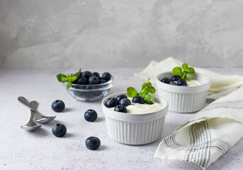 Milk yogurt with blueberries in white vases on a light table. Blueberries and mint leaves in the background. Dessert spoons on the table. Light grey background