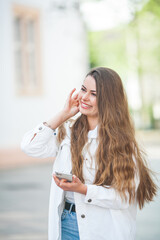 A young beautiful woman with long hair in comfortable light casual clothes listens to music using bluetooth headphones, walks in the center of a European city. A cute model is shopping. Spring.
