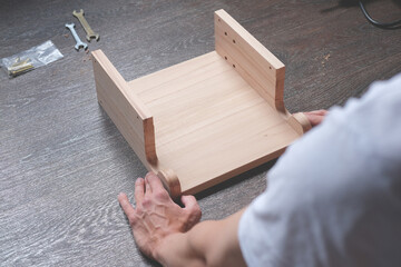 Man with his back in a white t shirt assembles a handcraft natural wooden furniture shelf next to some instruments and tools on floor at home