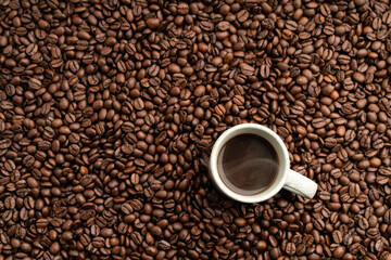 Small coffee cup filled up with hot coffee placed onto bed of roasted coffee beans flat lay view