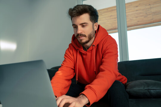 Young man wearing red hoodie remote working from home and having work conference video call. Sitting on sofa In living room, working remotely on online project.