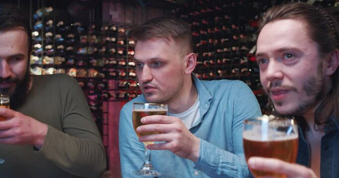 Caucasian handsome guys drinking beer in pub at night, talking and watching sport match. Mest males friends sipping drinks and cheering up for game on sport channel. Footbal fans hanging out in bar.