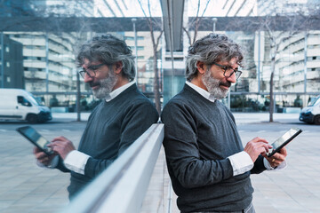 Portrait of contemporary senior man using digital tablet and browsing internet while standing against window, copy space