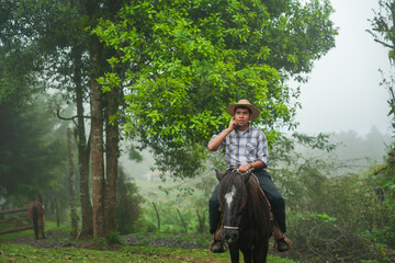 A young man talks on the phone while riding his horse through a foggy trail.
