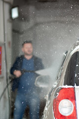 A man in a blue jacket and sunglasses washes his car at a self-service car wash.