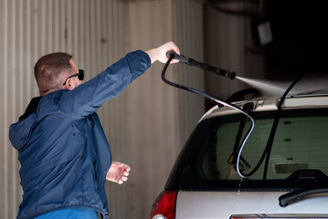 A man in a blue jacket and sunglasses washes his car at a self-service car wash.