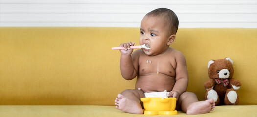 Adorable African baby newborn in diaper sitting on sofa with small bear doll trying to grab a spoon...