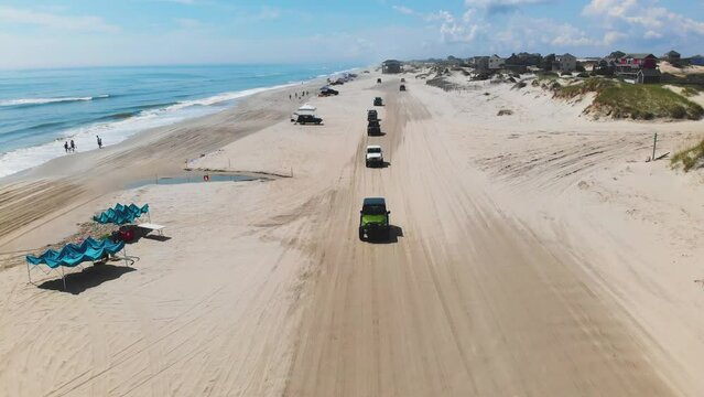 Hovering drone above multiple Jeeps passing by on Outer Banks Corolla 4x4 beach, houses can be seen inland.