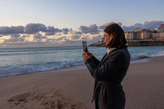 A girl photographs the sunset on the ocean beach in a coat.