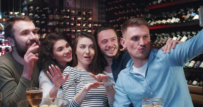 Loud cheerful company of Caucasian friends cheering in bar and taking selfie photo on smartphone. Happy men and women posing together for mobile phone camera. Drinking beer in pub.