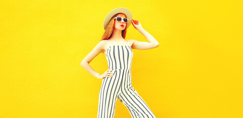 Stylish model woman wearing summer round straw hat, white striped jumpsuit posing on yellow background