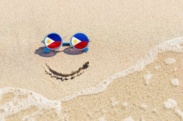 Fototapeta na wymiar A painted smile on the beach and sunglasses with the flag of the Philippines. The concept of a positive and successful holiday in the resort of the Philippines.