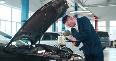 Caucasian male mechanic in uniform and goggles filling in document form about repairment in auto service. Young man fixing engine of vehicle and writing bill. Maintaining work concept.