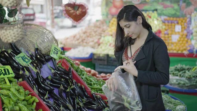 Young attractive woman shopping at the greengrocer choosing eggplant. She consumes fresh organic fruits for a healthy life. City life concept. Vegan food ingredients.