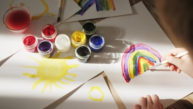Child paints a rainbow. The rainbow is drawn by a close-up of children's hands with multi colored paints on a white paper.