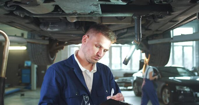 Caucasian male mechanic in uniform and googles repairing under car with wrecker in auto service. Young man engineer fixing engine and twisting spanner from underground. Maintaining concept.