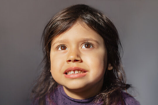 A Little Cute Girl With Brown Eyes Looks Up Hopefully, Asks For Sweets Or Needs Help. Hopeful Brown Hair Isolated On Gray Studio Background. Can I Please Concept.