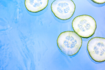 Cucumber slices in transparent water with droplets and bubbles on blue background. Trendy, abstract, summer hydration and refreshing background.