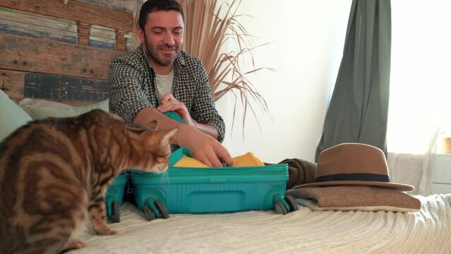 Man packs winter themed cyan bag for holiday. Vacation preparation, careful bag packing. Millennial generation Y travel blogger pack suitcase and petting cat. loft design room. 4k footage