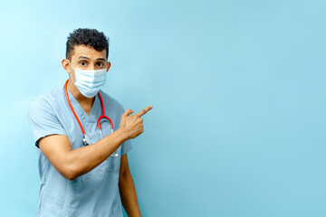 Hispanic man with face mask with hand points to something on a blue background.