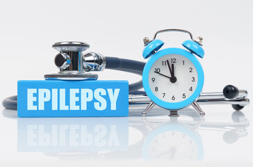 On a white surface there is an alarm clock, a stethoscope and a blue block with the inscription -...